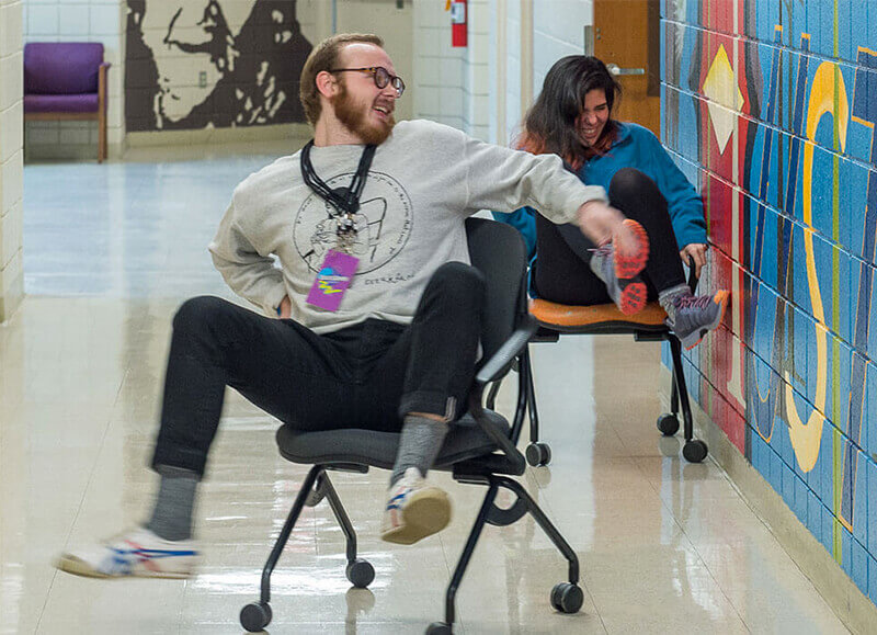 Students rolling on chairs down a hallway.